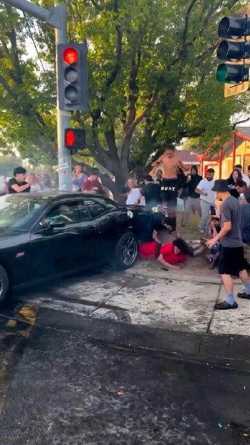 A black car performing donuts at an intersection has struck a group of people, causing several to fall to the ground. The scene is chaotic, with bystanders reacting in shock and some are trying to assist those who were hit. Smoke from the car's tires and a traffic light are visible.