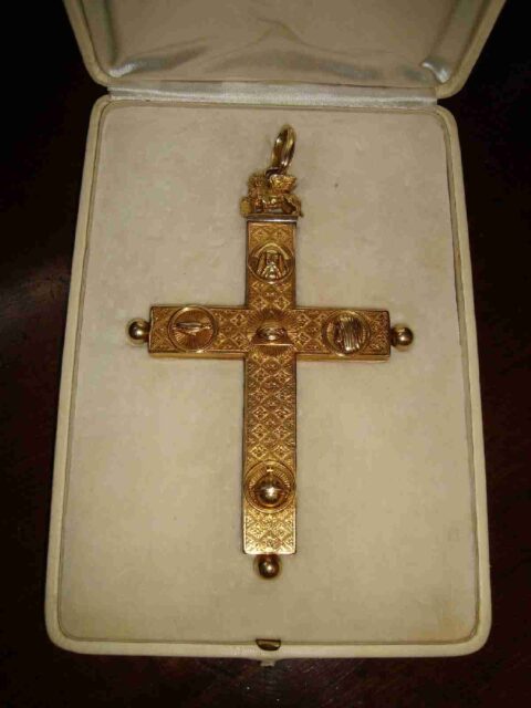 A gold cross, intricately designed and decorated with symbols. At the top of the cross, there’s a winged lion associated with Saint Mark, and below it, God as an old bearded patriarch. The center of the cross features the All-Seeing Eye, also known as the Trinity, encased within a radiant sunburst. On the left arm of the cross, there’s a dove facing towards the All-Seeing Eye. On the right arm of the cross, there's a head in profile, also facing the All-Seeing Eye. Finally, at the bottom of the cross, there's a globe or orb with rays extending outward. The cross is presented in a cream-colored cushioned case, underscoring its importance and the care taken in its preservation.