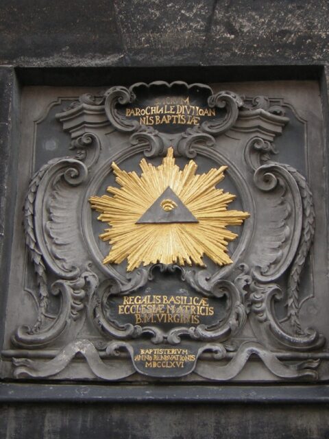 The All-Seeing Eye depicted on the gate of Aachen Cathedral in Germany. The eye is enclosed within a triangle, set against a background of golden rays and intricate gray stone carvings. Latin inscriptions are featured above and below the symbol, enhancing the ornate design of the gate.