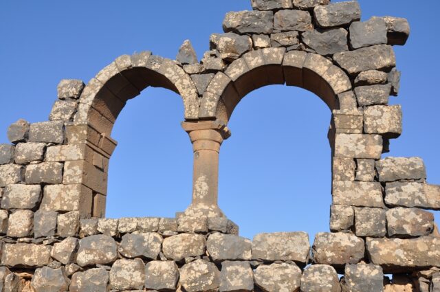 An ancient stone structure featuring two adjacent arched windows with a single central column between them. The stonework is weathered and uneven, with some stones missing or crumbling.