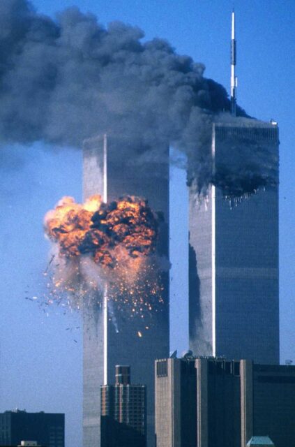 A photograph of the World Trade Center towers during the September 11, 2001 attacks. The North Tower (right) is emitting thick black smoke after having been struck by an airplane, and the South Tower (left) has just been struck by an airplane, resulting in a large explosion with flames and debris.