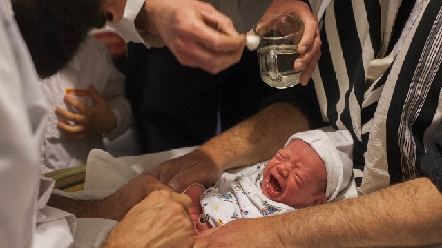 A crying infant being held by an adult while a religious figure performs the circumcision using a specialized knife. Someone is standing by holding a cotton ball and a glass of water.