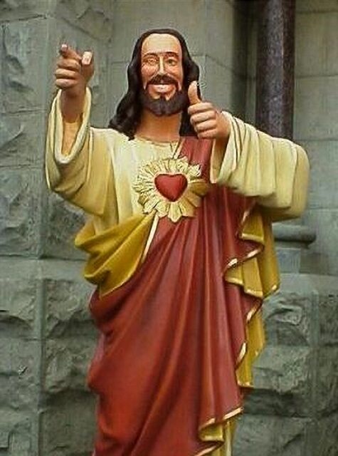 A statue of "Jesus Christ." The statue shows Jesus smiling broadly, giving a thumbs-up with one hand, and pointing with the other, with his Sacred Heart visible on his chest. The figure is depicted in bright colors, standing against a stone background.