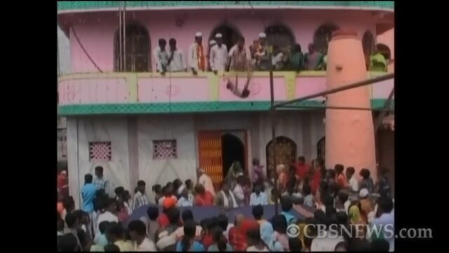 A crowd of people gathers around a two-story building, where a ritualistic baby-dropping ceremony is taking place. The event, captured in the image, shows a baby being dropped from the balcony of the building to be caught below. The image is sourced from CBS News.