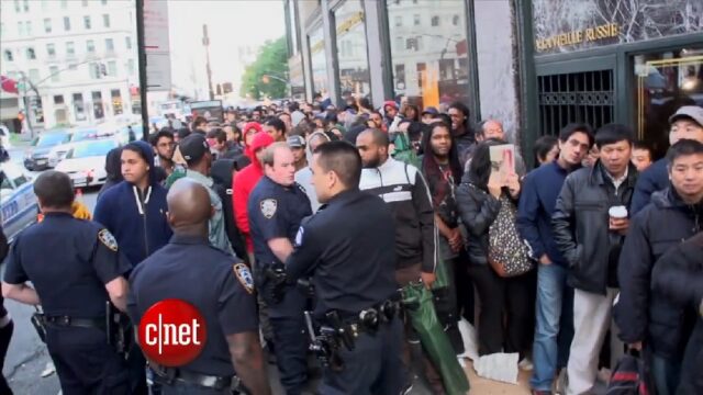 A large crowd of people is standing in line, waiting for the release of the Apple iPhone 6. Several police officers are present to maintain order. The scene is busy and energetic, taking place in Manhattan on Fifth Avenue. The CNET logo is visible, indicating that this is part of a news report.