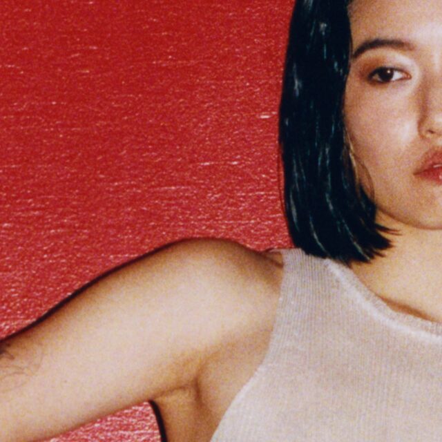 A close-up of a woman, with only the right side of her face visible. She has straight, dark hair and wears a sleeveless top against a red wall. Her expression is neutral, almost melancholic. This image conveys a minimalist aesthetic, emphasizing simplicity and color contrast.