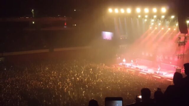 A large concert crowd is gathered in front of a brightly lit stage, where a band is performing. The audience is densely packed, with many people raising their hands and some holding up phones to record the event. The crowd is creating a mosh pit by running in circles, adding to the energetic and chaotic atmosphere.