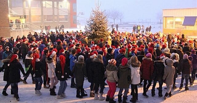 A large group of adolescents, many wearing Santa hats, hold hands and dance in circles around a Christmas tree outdoors. The scene is festive and snowy, with people bundled up in winter coats, enjoying the communal celebration. Buildings and other participants are visible in the background, contributing to the lively holiday atmosphere.