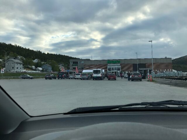 A view from inside a car showing a parking lot with several cars clustered near the entrance of a Europris store. The surrounding area appears relatively empty, with ample parking spaces farther from the entrance. The sky is cloudy, and a few people can be seen walking near the store.