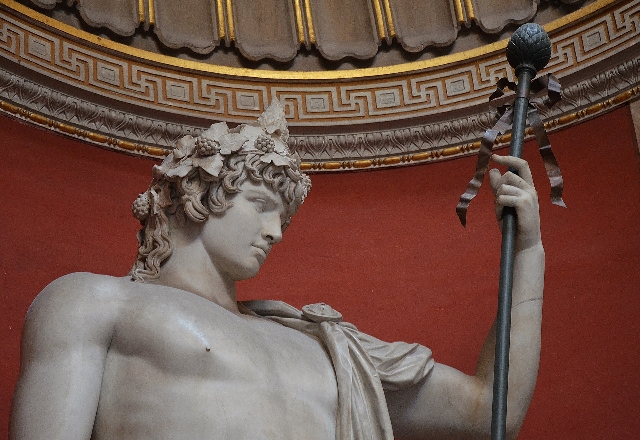 A colossal statue of Antinous depicted as Dionysus, adorned with a laurel wreath and holding a thyrsus (a staff topped with a pine cone) in his left hand. The background features ornate architectural details, enhancing the classical and majestic appearance of the sculpture.