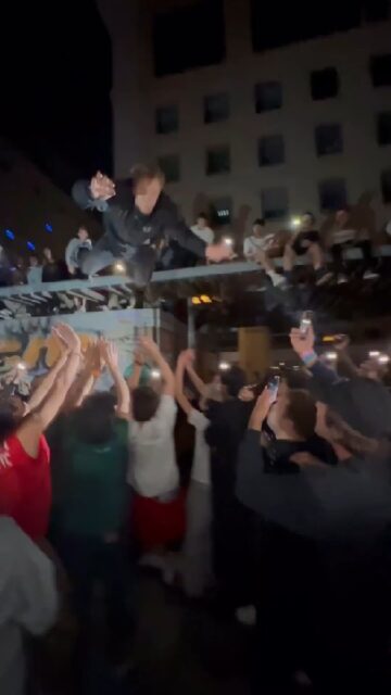 A person is mid-air, having jumped from a platform above a crowd at night. The crowd below is reaching up, seemingly ready to catch the jumper. The scene is brightly illuminated by a camera flash, with many people in the crowd holding up their phones, capturing the moment.