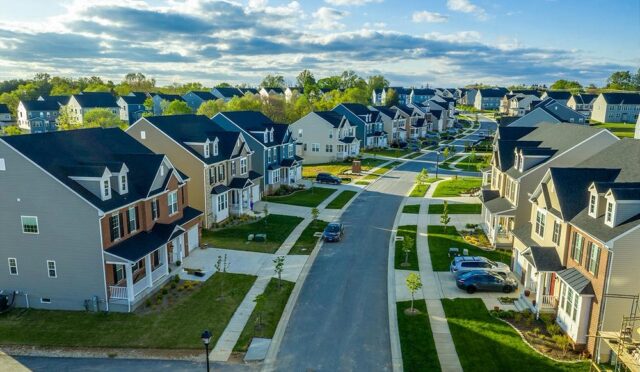 A rich suburban neighborhood with rows of densely packed, massive modern houses, featuring well-maintained lawns, driveways, and sidewalks. The homes are large and similar in design, indicating a planned community of high-value properties.