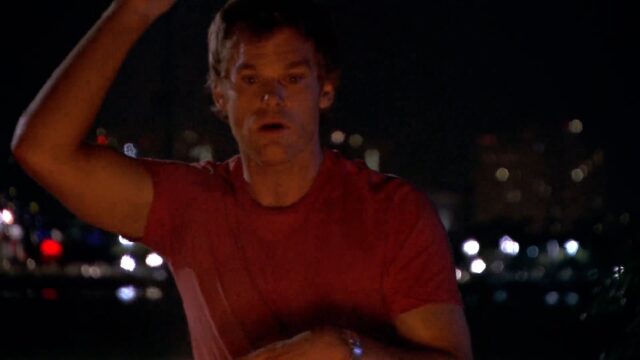 Dexter Morgan, wearing a red shirt, stands behind a campfire at night, telling a story while raising his right hand getting ready to smash it down. The dark background is illuminated by distant city lights.