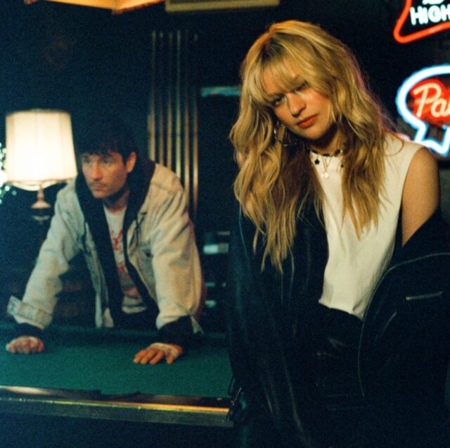 A woman and a man in a dimly lit bar setting. The woman's expression appears distant and contemplative, while the man leans against a pool table in the background, creating a sense of tension or reflection.