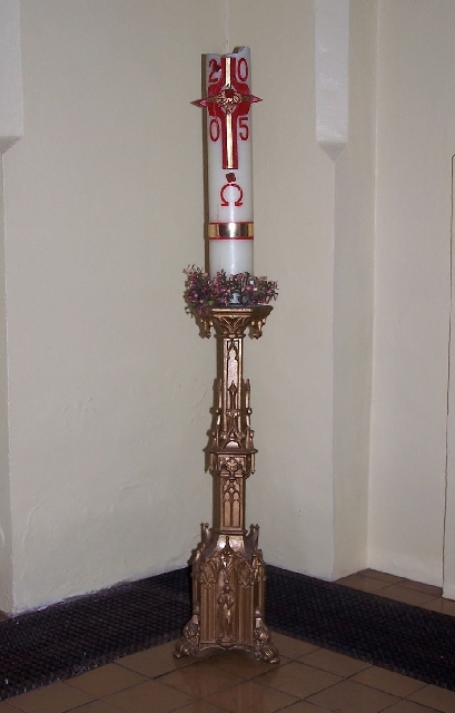 A tall Paschal candle with red symbols for the year 2005, the Omega symbol under the Christian cross, standing on an ornate gold stand in a church. There was a red Alpha symbol above the cross before it melted away.