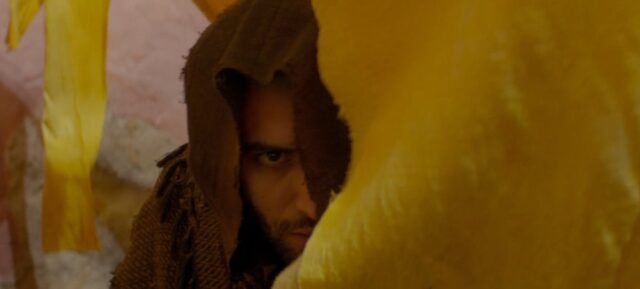 Jafar, dressed in a brown cloak, looks intently from behind yellow fabric, targeting Aladdin as he prepares to steal the magical lamp from him. Only the right side of his face is visible, as the left side is covered by his hood.