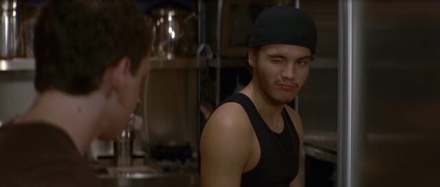 A scene from "Alpha Dog" showing Johnny Truelove, wearing a black tank top and backwards cap, winking at Zack. Johnny is standing in a kitchen, giving a sly expression, hinting at his deceptive intentions.