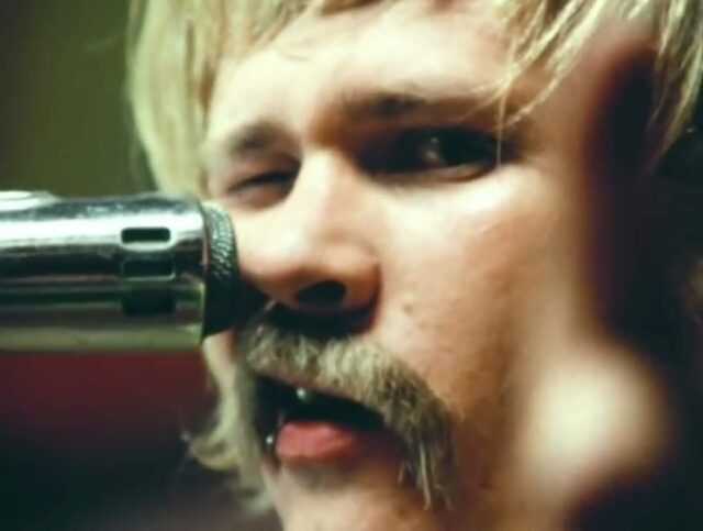 Tom DeLonge in the music video "Blink-182 - First Date," winking at the camera while singing into a microphone, sporting a retro mustache and blonde hair.