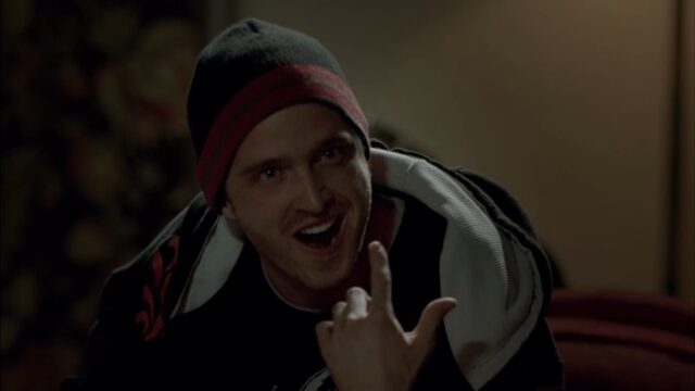 A scene from "Breaking Bad," where Jesse Pinkman, is pointing at his left eye, which is bruised and swollen (a black eye), while smiling and wearing a black beanie with a red stripe.