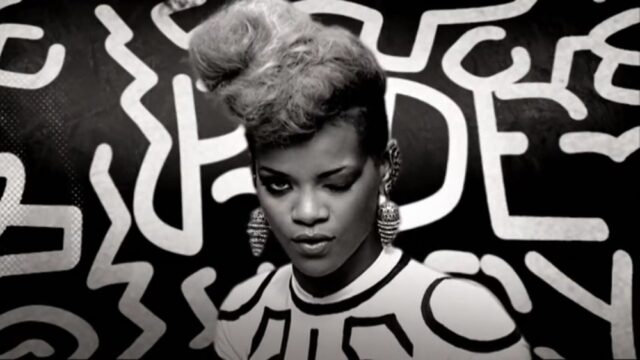 Rihanna winks at the camera in the music video for her song "Rude Boy," standing in front of a bold, graphic black-and-white background.