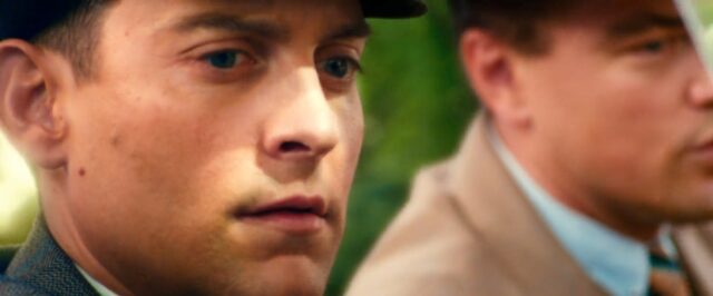 A scene from "The Great Gatsby" featuring the character Nick Carraway in close-up, with a thoughtful expression. In the background, the character Jay Gatsby is partially visible, out of focus, with only one eye in frame. Jay is steering as they drive in a roofless car.