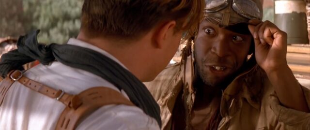 A scene from "The Mummy Returns" featuring the character Izzy Button, wearing aviator goggles on his head and speaking to the main character Richard O'Connell. He is lifting his eye patch, revealing that his eye is perfectly fine.