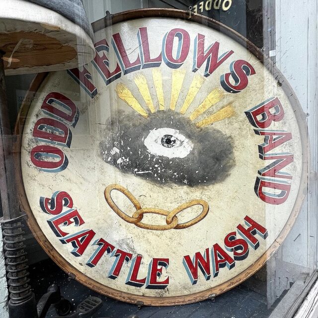 A vintage drum displayed at the Odd Fellows Hall in Capitol Hill, Seattle, Washington, featuring the Eye of God surrounded by rays of light emerging from a cloud. The text "Odd Fellows Band Seattle Wash" is painted around the drum, with an image of three linked golden chains beneath the eye.