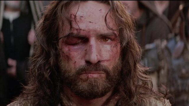 A man with long hair and a beard, his face bruised and bloodied, particularly around one swollen eye.