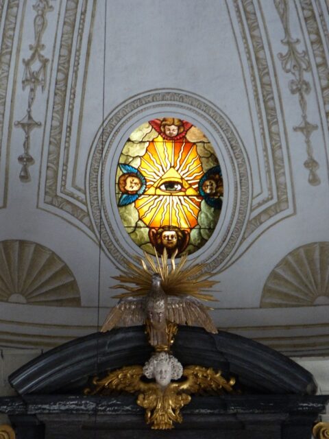 A stained glass depiction of the Eye of Providence above the altar in the Church of St. Leodegar in Lucerne, Switzerland. The eye is enclosed in a triangle with rays of light emanating from it, and is surrounded by cherubic faces.