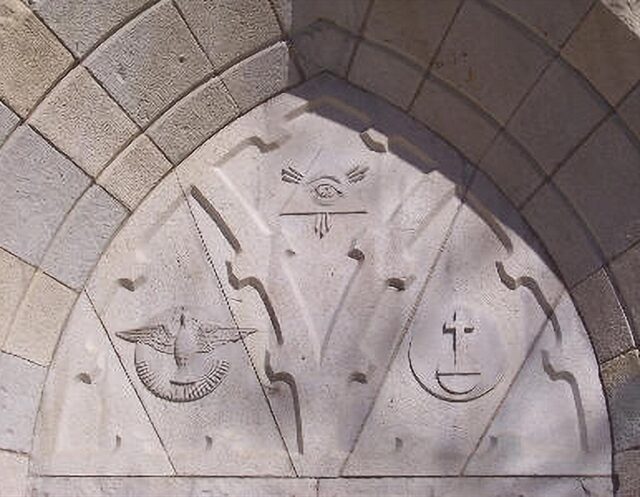 A stone relief above an arched entrance at the Church History Museum in Salt Lake City. The relief features three symbolic elements: the Eye of Providence at the top, a bird representing the Holy Spirit on the left, and a cross with a chalice-like symbol on the right. These symbols are carved into the stone, each enclosed within a triangular frame, signifying aspects of Christian theology.