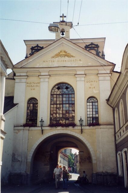 The Eye of Providence depicted on the facade of the Gate of Dawn in Vilnius, Lithuania. The symbol is carved above the entrance, surrounded by rays of light. The historic gate features arched windows and a passageway beneath, with people walking through the arch. The inscription "Mater Misericordiae" is visible under the Eye of Providence.