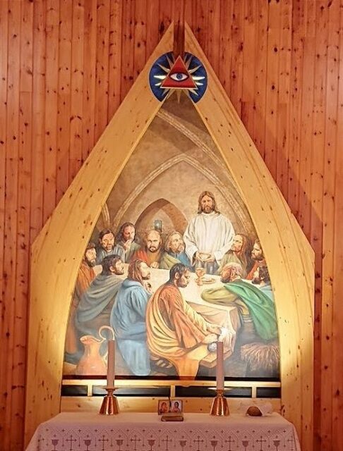 The altar of Arnøy Church in Norway featuring a painting of the Last Supper beneath a triangular symbol of the Eye of Providence. The eye is depicted within a red triangle, surrounded by rays of light against a blue background, representing divine oversight over the scene of Jesus and his disciples at the table.