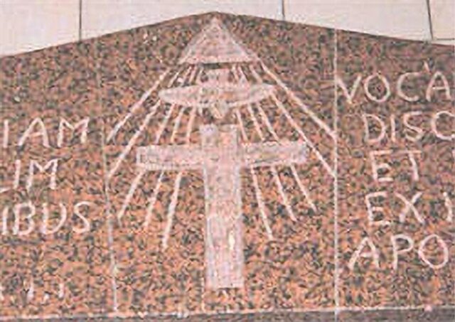 A depiction of the Eye of Providence in the Church of the Annunciation in Nazareth, Palestine. The Eye of Providence, symbolizing God, is positioned above a bird representing the Holy Spirit, which in turn is above a cross symbolizing Jesus. Rays of light emanate from the eye, and Latin text is inscribed on either side of the image, all carved on a brown stone background.