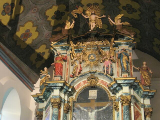 The elaborate altarpiece in Our Lady's Church in Trondheim, Norway, featuring the Eye of Providence at the top within a triangle, surrounded by golden rays and flanked by angels. Above the eye, a standing figure of Jesus Christ is depicted with his arms outstretched, as if in a gesture of triumph or invitation. Statues of saints and other religious figures are positioned around the ornate structure.