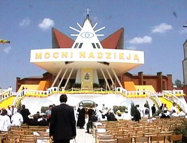 A Catholic church in Zamość, Poland, featuring a large Eye of Providence symbol at the top of its modern triangular structure. The church is adorned with the words "MOCNI NADZIEJĄ," which translates to "Strong in Hope," and is surrounded by chairs and attendees gathered for a religious event.