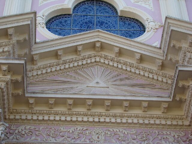 The Eye of Providence depicted on the exterior of a cathedral in Salta, Argentina. The symbol is carved within a triangular pediment above the entrance, with rays of light extending outward. The detailed architectural decorations complement the pink and white facade of the cathedral.