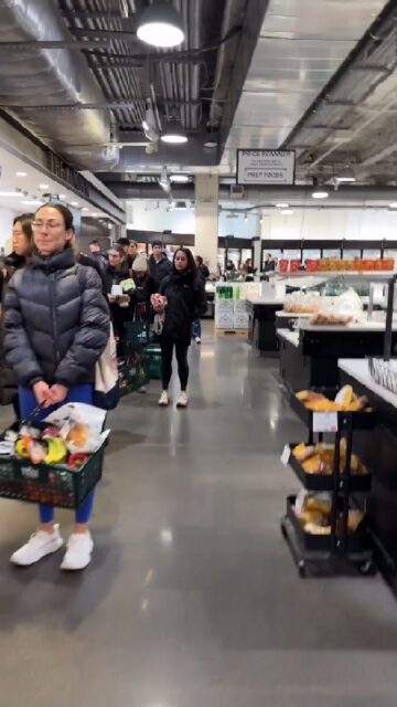 A long line of customers with shopping baskets wait to check out at a grocery store. The line stretches through the store, with people standing patiently, holding their items. The store has a modern industrial design with exposed pipes on the ceiling and bright lighting.