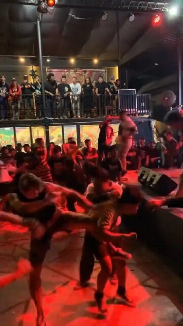 A group of people engaged in hardcore dancing at a concert, performing energetic and aggressive moves like spin kicks and windmills near the stage. One man is bendig over as he is being kicked in the stomach. The scene is illuminated by red stage lights, with an audience watching from both the floor and a balcony area in the background. The atmosphere is intense and dynamic, reflecting the high energy of the music.