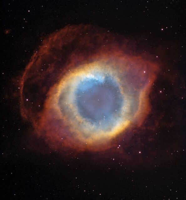 A detailed image of the Helix Nebula, a colorful cosmic formation resembling an eye. The nebula displays a vibrant blue center surrounded by layers of orange, red, and brown gases, set against the blackness of space dotted with small stars.