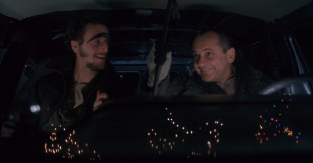 Two characters from the movie "Home Alone," Harry and Marv, sit in a van at night, with Christmas lights reflecting on the windshield. Both hold a crowbar each and share a mischievous smile.