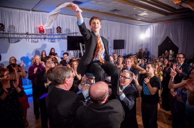 A young boy is lifted on a chair during the Hora dance at his Bar Mitzvah celebration, surrounded by smiling and clapping guests.