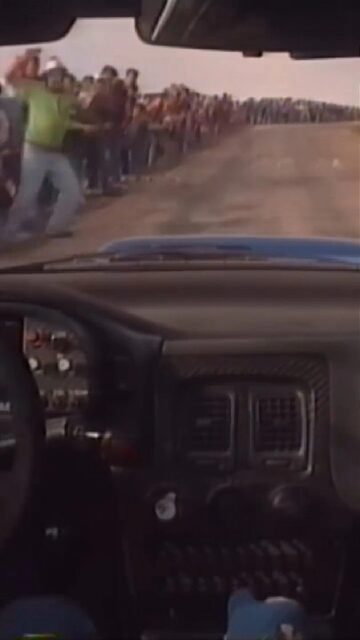 A view from inside a rally car, showing a dirt track ahead. A tight crowd of spectators are standing dangerously close on the side of the track, creating a narrow path for the rally car.