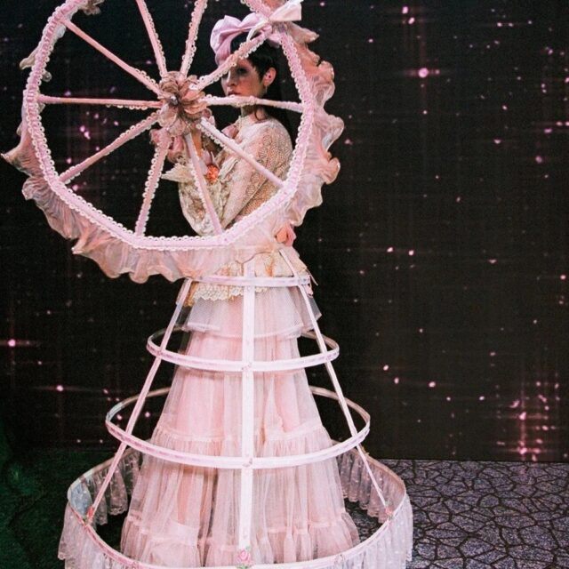 Melanie Martinez dressed entirely in pink, wearing a hoop skirt with a cage-like structure and holding a ferris-wheel-like pink umbrella in front of her face, covering her right eye. The outfit blends avant-garde fashion with whimsical elements, creating a surreal and artistic look.