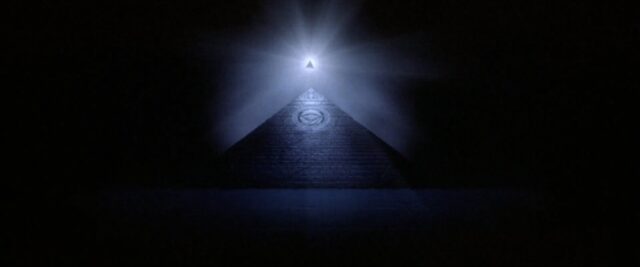 An illuminated triangle floating above a pyramid in the darkness, with light radiating from the triangle. A circular design resembling the All-Seeing Eye is visible on the pyramid's surface, emphasizing Illuminati symbolism.