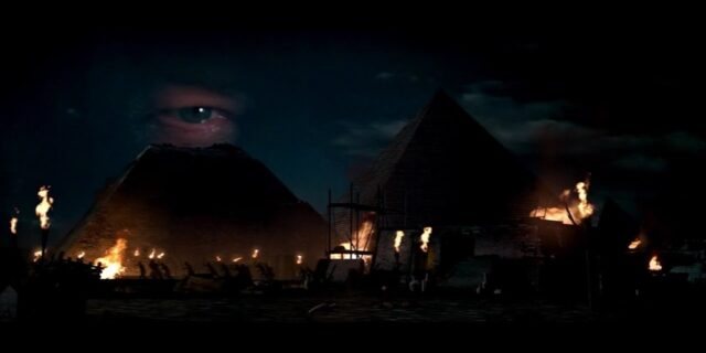 The image shows the right eye of John Adams Gates, prominently hovering above the pyramids of Giza at night. The eye is visible above a pyramid that is missing its top. The scene is illuminated by torches, with some areas of the pyramids and surroundings glowing in the firelight.