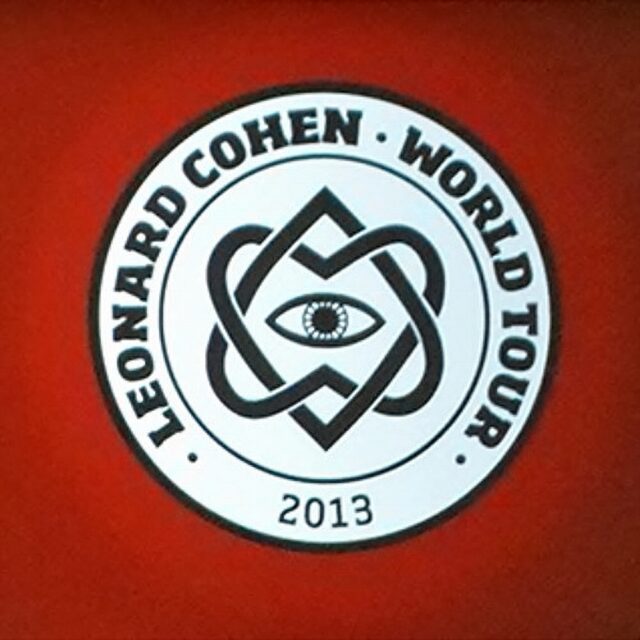 The logo for Leonard Cohen's 2013 World Tour, featuring the symbol of the Unified Heart—a design of two interlocking hearts forming a star-like shape with an eye in the center. The text "Leonard Cohen World Tour 2013" surrounds the symbol on a circular badge against a red background.