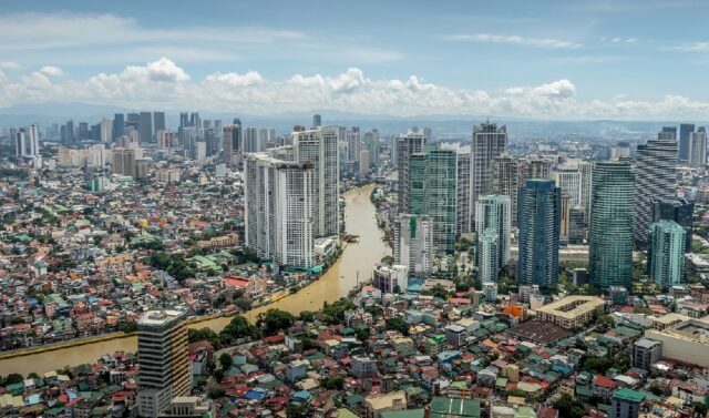 Aerial view of Manila in the Philippines, showcasing a densely populated urban area with a mix of high-rise buildings and residential neighborhoods, and a dirty river flowing through the city. The skyline features a combination of modern skyscrapers and older structures, illustrating the city's diverse architecture and high population density.