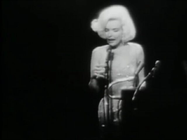 A black and white image of Marilyn Monroe is standing in the spotlight while singing "Happy Birthday" to President John F. Kennedy at the celebration held in his honor in 1962. She is dressed in a sparkling, form-fitting gown, standing in front of a microphone on a dimly lit stage.