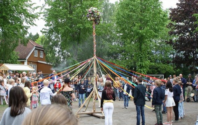 A group of people, including children and adults, are gathered around a decorated maypole with colorful ribbons attached. Children are holding the ribbons and appear to be preparing for or engaging in a dance around the maypole. The scene takes place outdoors, with trees and houses in the background, during a traditional Midsummer celebration.