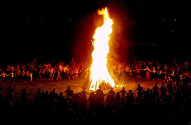 A large bonfire surrounded by people holding hands and dancing in a circle at night. The scene is illuminated by the flames, highlighting the festive and communal atmosphere.